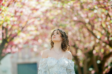 Beautiful young blonde in light romantic dress. Portrait of young woman near blooming sakura tree in a European city. Warm bright spring.