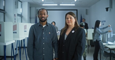 Portrait of multi ethnic couple, United States of America elections voters. Family stand in a modern polling station, pose, smile and look at camera. Background with voting booths. Civic duty concept.
