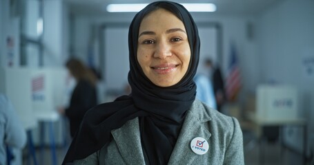 Portrait of Arabic woman, United States of America elections voter. Woman stands in a modern polling station, poses, smiles and looks at camera. Background with voting booths. Concept of civic duty.