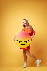 Social network concept. Happy young woman holding yellow angry emoji face. Colorful studio portrait.