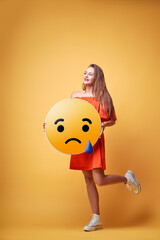 Social network concept. Happy young woman holding yellow sad emoji face. Colorful studio portrait.