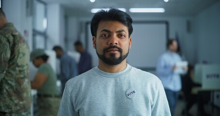 Man stands in a modern polling station, poses and looks at camera. Portrait of Indian man, United States of America elections voter. Background with voting booths. Civic duty and patriotism concept.