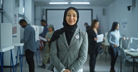 Woman stands in a modern polling station, poses, smiles and looks at camera. Portrait of Arabic woman, United States of America elections voter. Background with voting booths. Concept of civic duty.