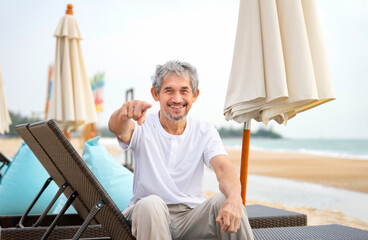 playful elderly man sitting on beach chair,smiling and pointing to camera,happy senior male enjoy holiday time on the beach