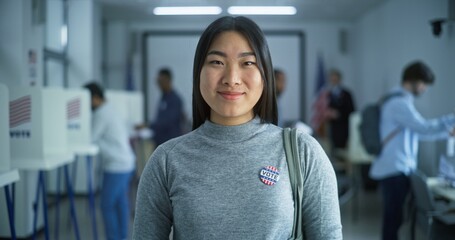 Portrait of Asian woman, United States of America elections voter. Woman with badge stands in modern polling station, poses, smiles, looks at camera. Background with voting booths. Civic duty concept.