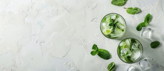 On a white concrete table, there are two highball glasses containing a traditional Mojito cocktail, made with rum, spearmint, lime juice, soda water, and ice, seen from above with space for text. - obrazy, fototapety, plakaty