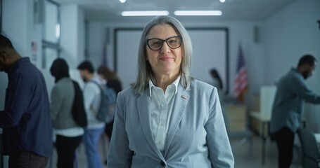 Portrait of mature woman, United States of America elections voter. Businesswoman stands in a modern polling station, poses and looks at camera. Background with voting booths. Concept of civic duty.