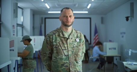 Portrait of male soldier, United States of America elections voter. Man in camouflage uniform stands in a modern polling station and looks at camera. Background with voting booths. Civic duty concept.