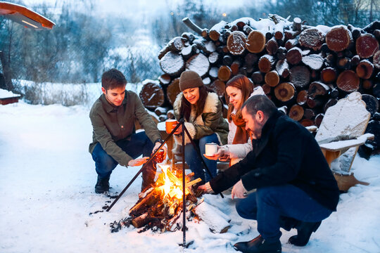 Group of friends gathering around bonfire in backyard, drinking tea and warming hands. Two happy couples relaxing and enjoying winter season while sitting around fire. Outdoor winter entertaining