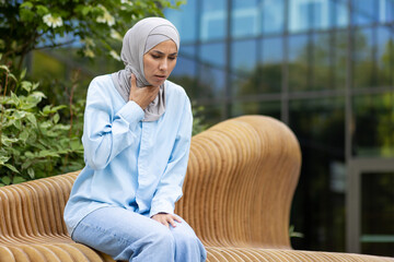 A young Muslim woman wearing a hijab looks uneasy as she sits on a wicker bench outside a modern...