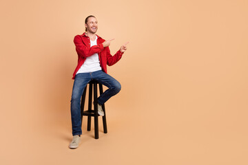 Fototapeta na wymiar Full length portrait of nice young man sit chair direct fingers empty space wear red shirt isolated on beige color background