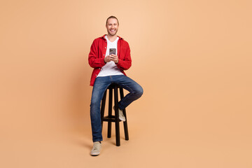 Full length portrait of nice young man sit stool hold phone wear red shirt isolated on beige color...