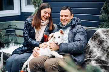 Young couple hugs their white cute pet dog. Man and woman at new year fair, drinking mulled red wine