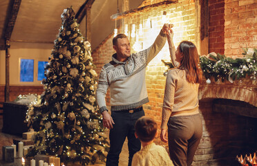 Happy family parents with two kids dancing together near fireplace and xmas tree in cozy...
