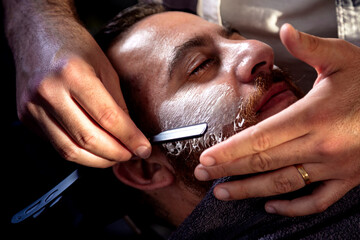 Barbershop. Barber shaving bearded male with a sharp razor. - obrazy, fototapety, plakaty