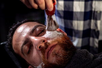 Professional hairdresser applying foam on customer's cheeks for shaving. - obrazy, fototapety, plakaty