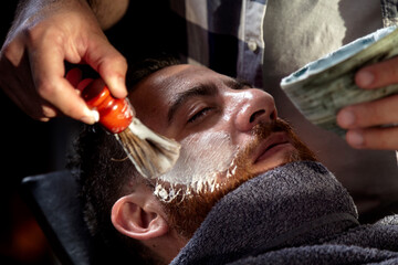 Working in Barbershop. Professional Barber applying foam on customer's cheeks for shaving. - obrazy, fototapety, plakaty
