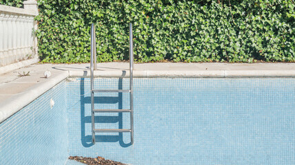 Blue tiled walls and steps of an empty dry swimming pool. Background wall of green leaves. Vacation low season. Square pattern of blue ceramic tiles, mosaic. Summer recreation in Spain - obrazy, fototapety, plakaty
