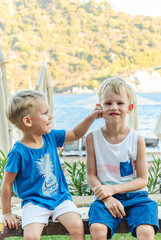 Two little boys are sitting on a bench near  beach laughing and engaging playfully on background of sea and hills and folded beach umbrellas