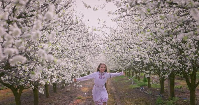 Beautiful smiling girl in white ethnic folklore embroidery slavic dress running outdoor under blooming spring trees in a garden