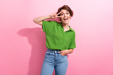 Photo of pretty excited young woman dressed green shirt showing v-sign cover eye isolated pink...