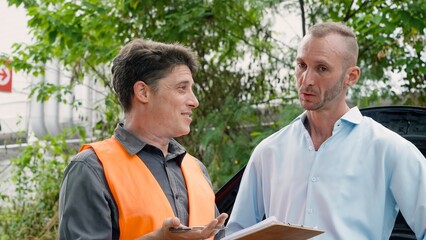 Two men discussing, one in orange vest holding clipboard, outdoors, plants in background. Concept of taking care of car insurance after an accident.