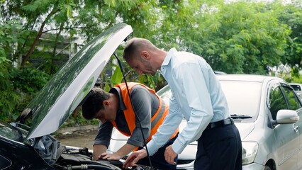 Middle-aged male in light blue shirt peers into car engine while mechanic in orange vest works,...