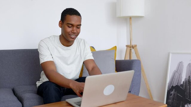 Happy young adult African man using laptop sitting on couch at apartment. Technology and education concept.
