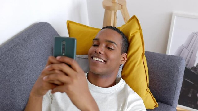Young adult man using mobile phone on the sofa. Happy millennial male chatting on smartphone app relaxing at home. Technology lifestyle concept.