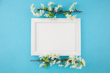 Top view photo of white wooden photo frame and branch with flowers on pastel blue background