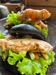A plate of food with a black bun and lettuce. The bun is topped with a sauce and a piece of salted egg soft shell crab. Selective focus.