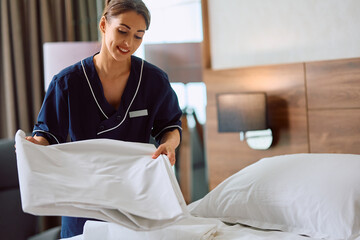 Happy chambermaid changing bedding in  hotel room.