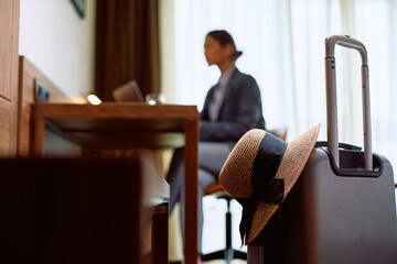 Sun hat and suitcase in hotel room with businesswoman in  background.