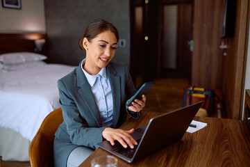 Smiling businesswoman using cell phone while working on laptop in hotel room.