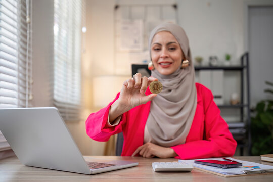 A female Muslim investor who trades in cryptocurrencies sits looking at stock charts and studying Bitcoin.