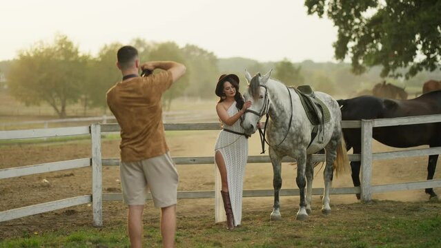 Young photographer takes picture of elegant girl on ranch with horse.