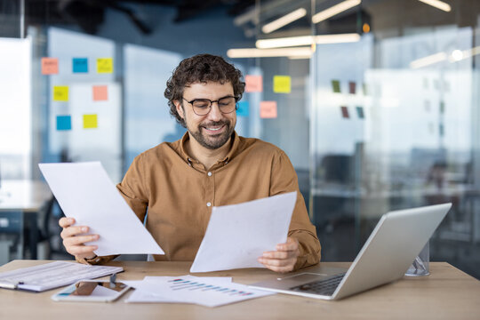 Happy businessman checking documents with records while sitting by table with portable computer and tablet. Satisfied person calculating financial indicators and comparing expenses and income. - Powered by Adobe