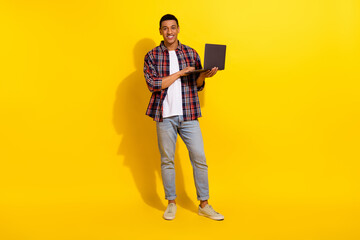 Full size photo of intelligent good mood man dressed plaid shirt denim pants hold laptop isolated on vivid yellow color background