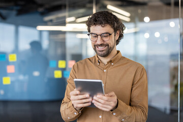 Smiling man in spectacles holding digital tablet while standing in corporate building with glassy...
