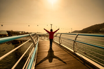 Traveler woman wear red clothes and raising arm standing on Songdo Skywalk at sunset in Busan, South Gyeongsang Province, South Korea.