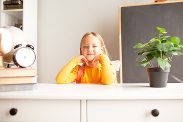 a little girl 6-7 years old is getting ready for school, the girl is dressed in a yellow long sleeve and sitting at a desk against the backdrop of desk