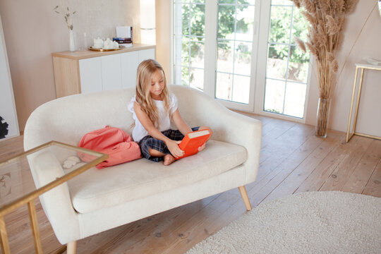 a little girl 6-7 years old is getting ready for school, the girl is dressed in a light-colored top and a plaid skirt.  The kid lsit on the sofa  with a backpack and a book