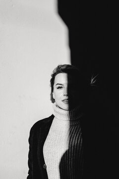 A woman stands before a wall, her gaze fixed on the camera with a serious expression. Clad in a black jacket and white sweater, the image exudes a moody and dramatic ambiance.