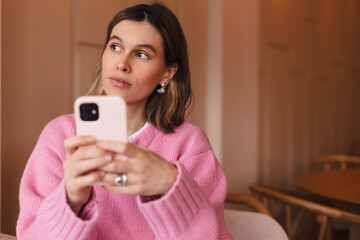 Confused puzzled woman in casual clothes, sits in cafe, holds a smartphone in her hand, looks...