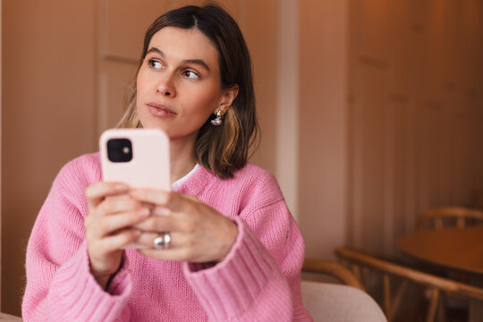 Confused puzzled woman in casual clothes, sits in cafe, holds a smartphone in her hand, looks questioningly, doubt. Girl think about solution, answer.