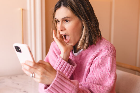 Joyful blonde woman in pink sweater playing video game on mobile phone, reading good news, celebrating winning lottery, feeling so happy and cheerful. Wow, win emotions, happy scream, hand near face.