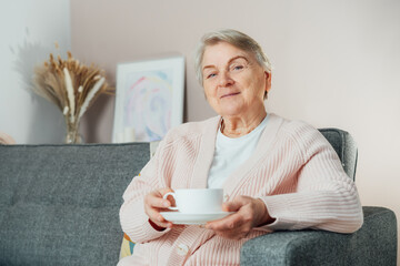 Smiling mature woman holding cup of tea, relaxing at home, positive senior female sitting on couch...