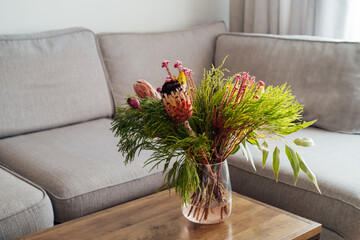 Vase with exotic protea flowers bouquet on coffee table with blurred background of modern cozy light living room with gray couch sofa. Open space home interior design. Elegant home decor. Copy space.