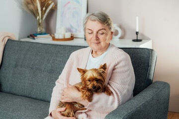 Elderly retired senior woman with wrinkles smiling while embracing her Yorkshire terriers dog pet and relaxing with pet on sofa at home. Best friend. Enjoying retirement lifestyle.