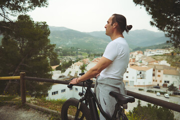 Side view of a male cyclist in white t-shirt, having a break, standing by his electric bike,...